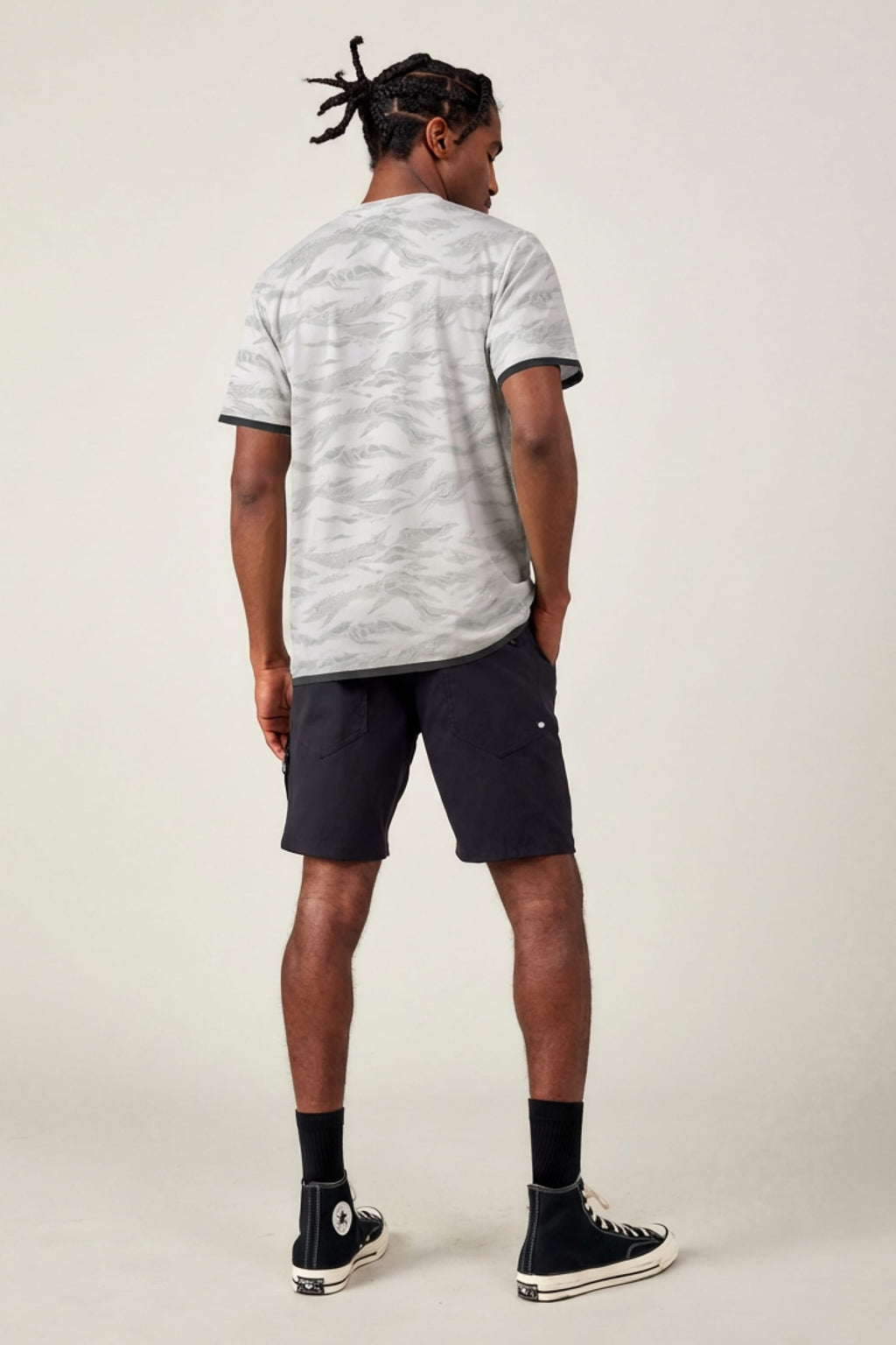 A man with short twisted hair faces away, wearing the 686 Men's Let's Go Tech Tee by 686—a light gray patterned breathable shirt—paired with black knee-length shorts, black crew socks, and black high-top sneakers against a plain light background.