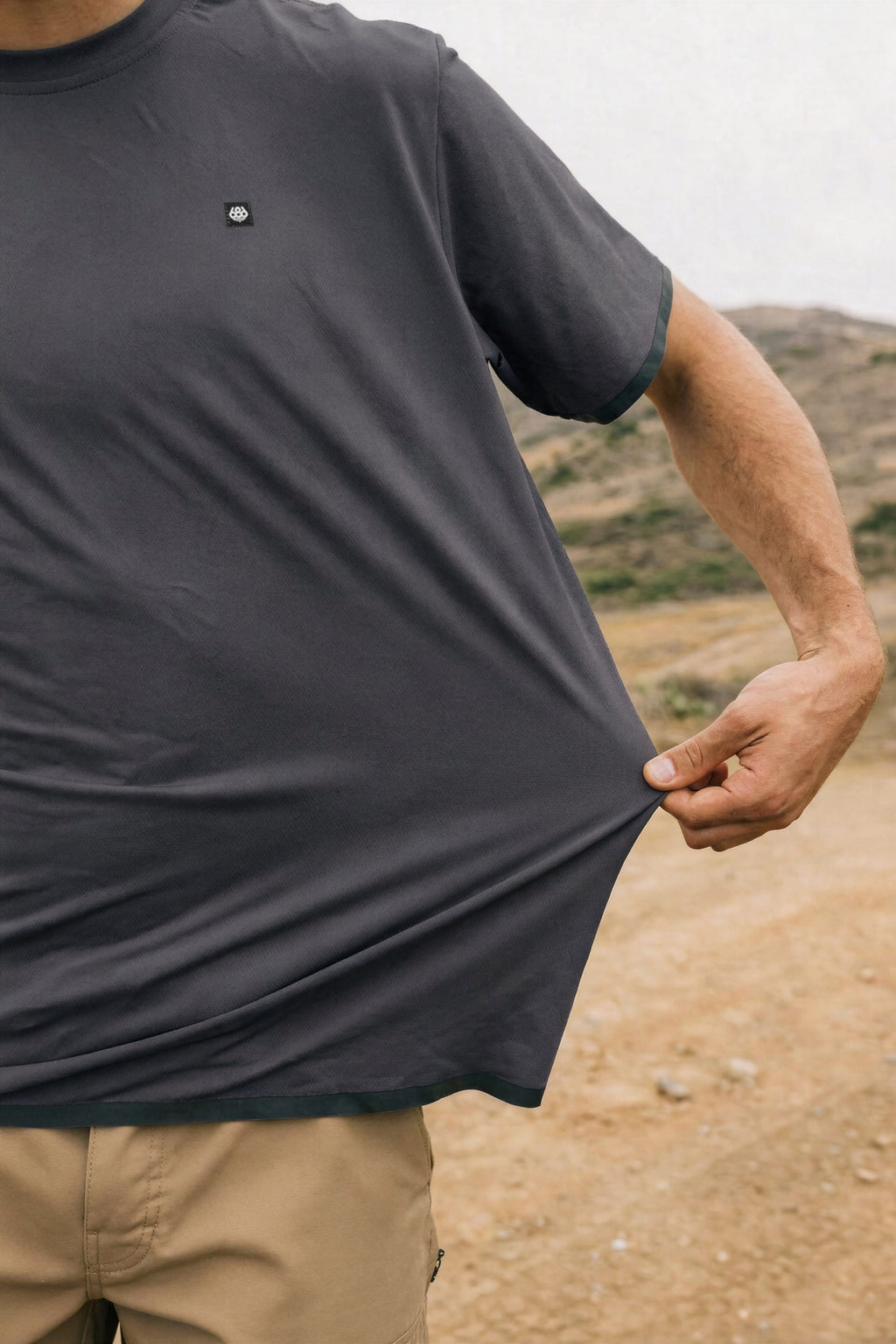 A person outdoors pinches and stretches the bottom of their loose, dark gray 686 Men's Let's Go Tech Tee; only their torso and arm are visible.