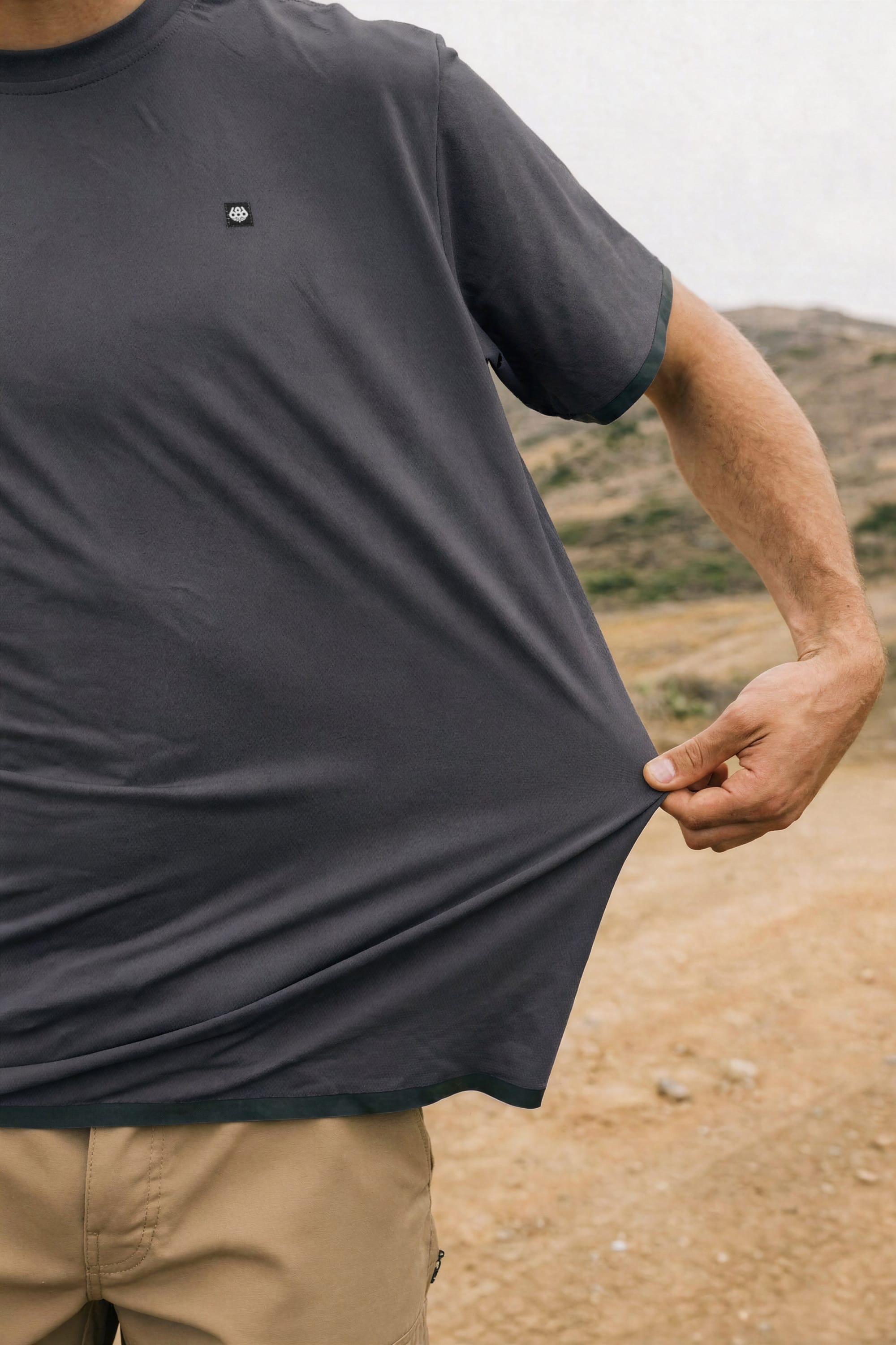 A person outdoors pinches and stretches the bottom of their loose, dark gray 686 Men's Let's Go Tech Tee; only their torso and arm are visible.