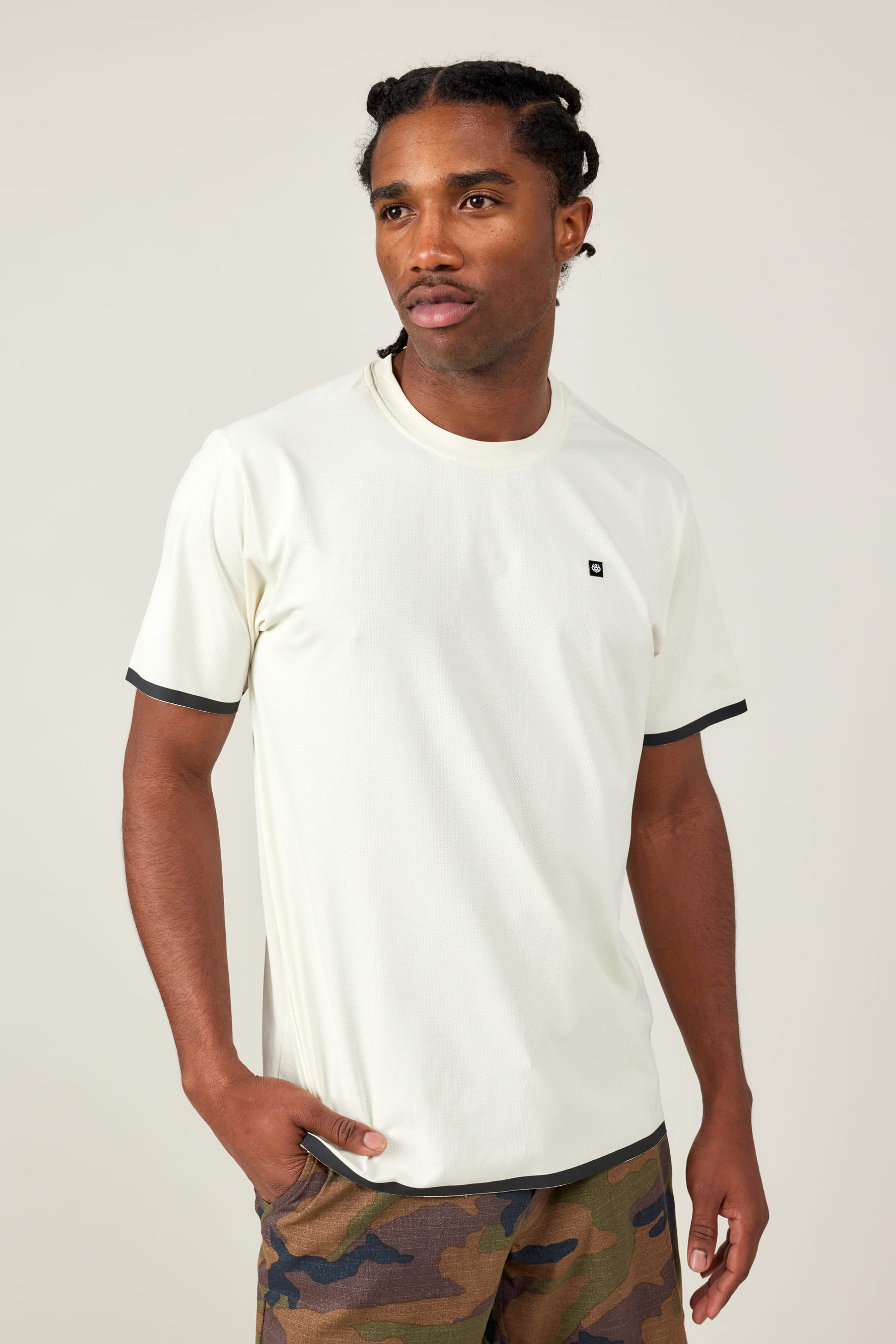 A man stands against a light background, wearing the 686 Men's Let's Go Tech Tee—a breathable, cream-colored shirt with black trim—and camo pants, looking off to the side with one hand in his pocket. Perfect for spring and summer adventures.