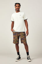 A man with braided hair stands against a plain background in the 686 Men's Let's Go Tech Tee, paired with camouflage shorts, black socks, and black high-top sneakers—a look blending 686’s effortless style and comfort for spring-summer adventures. One hand rests in his pocket.