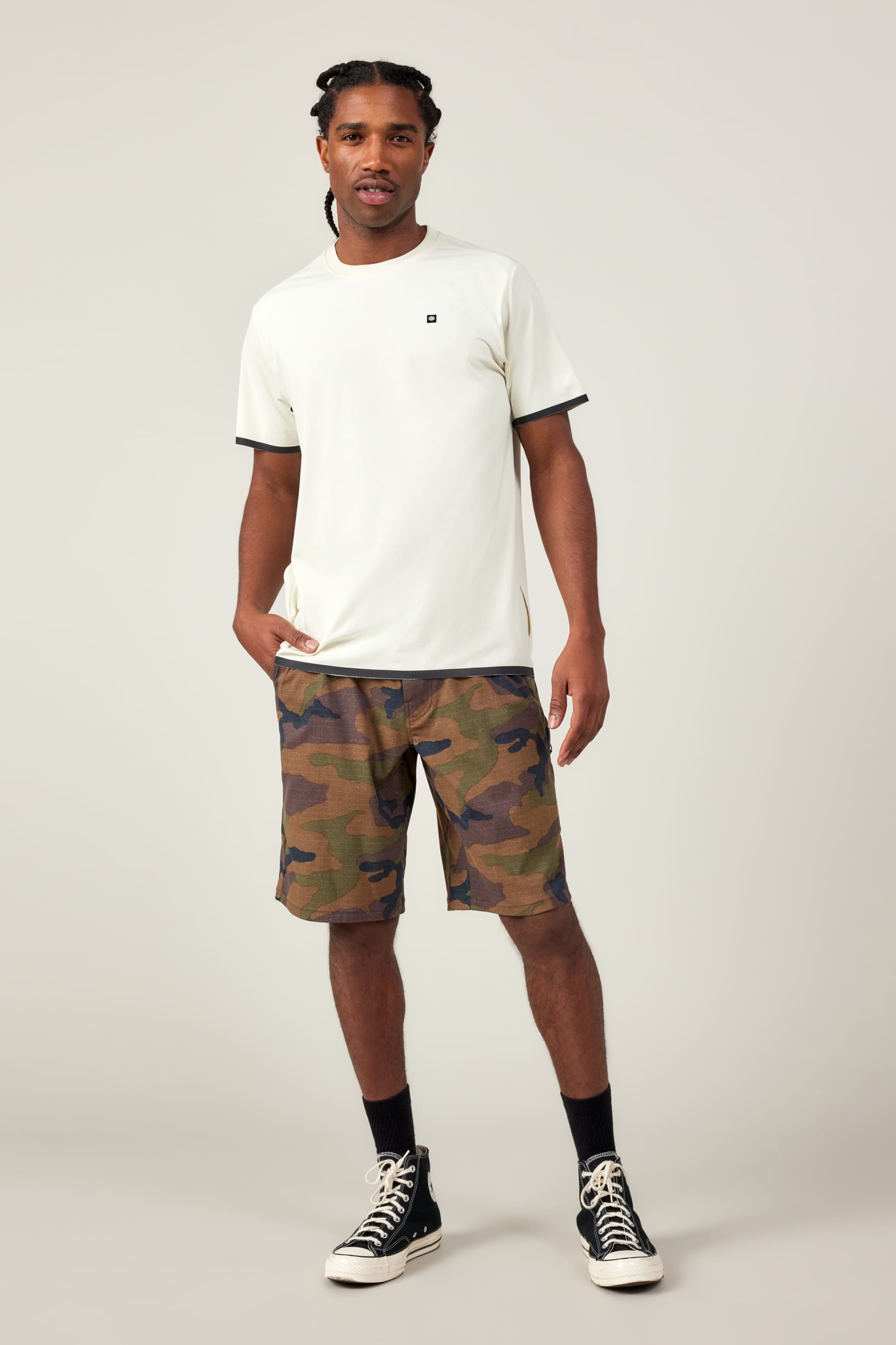 A man with braided hair stands against a plain background in the 686 Men's Let's Go Tech Tee, paired with camouflage shorts, black socks, and black high-top sneakers—a look blending 686’s effortless style and comfort for spring-summer adventures. One hand rests in his pocket.