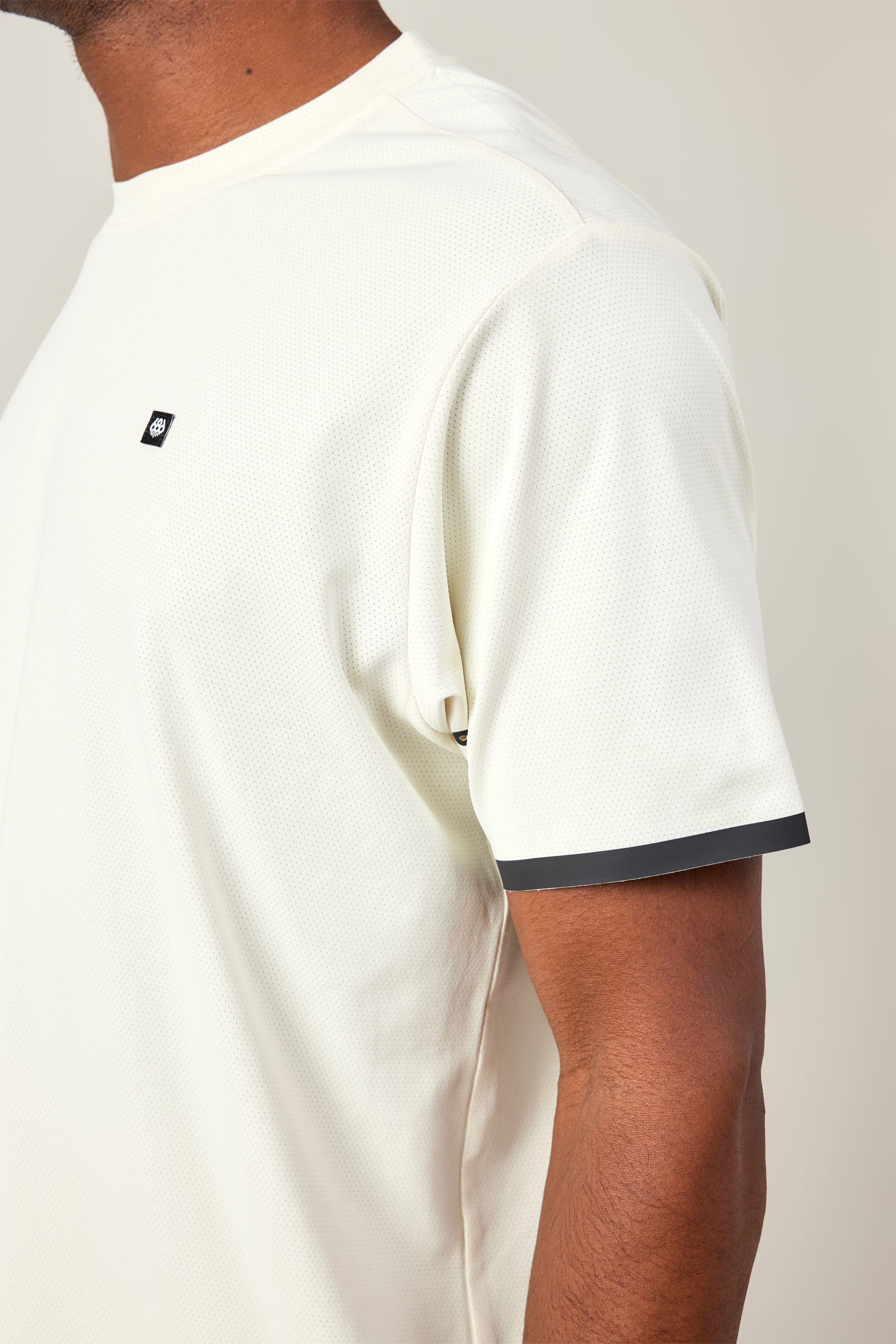 A person wears the 686 Men's Let's Go Tech Tee—a lightweight, off-white short sleeve shirt with a small black chest logo and black sleeve trim—ideal for spring and summer adventures. Shown from shoulders to mid-arm. Brand: 686.