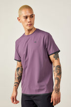 A man with short blond hair and tattooed arms wears the 686 Men's Let's Go Tech Tee in mauve and black pants, ready for spring-summer adventure. He looks to his left against a plain light background.