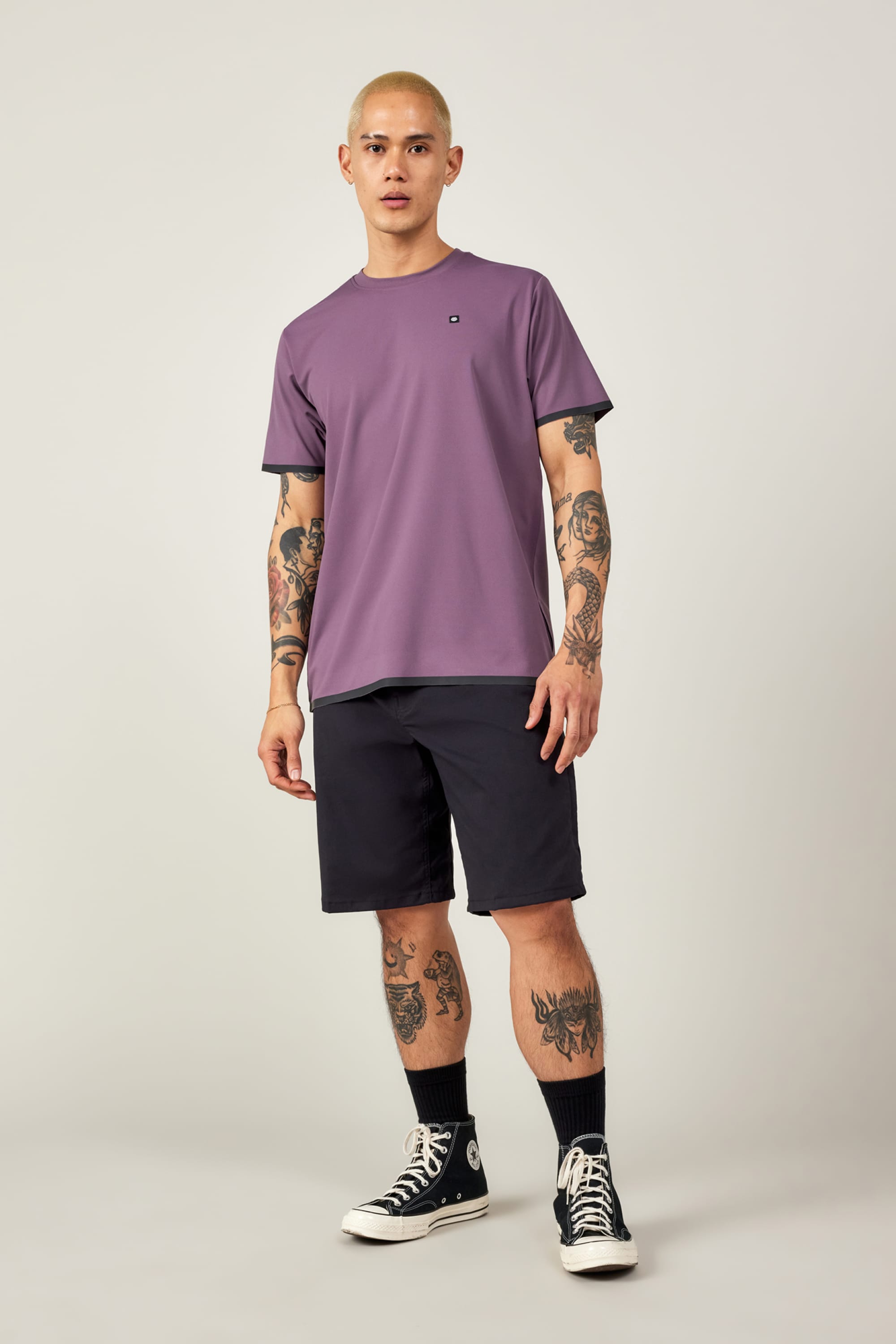 A man with short blonde hair and tattoos wears the 686 Men's Let's Go Tech Tee in purple, paired with black shorts, high black socks, and black Converse sneakers, standing against a plain light background.