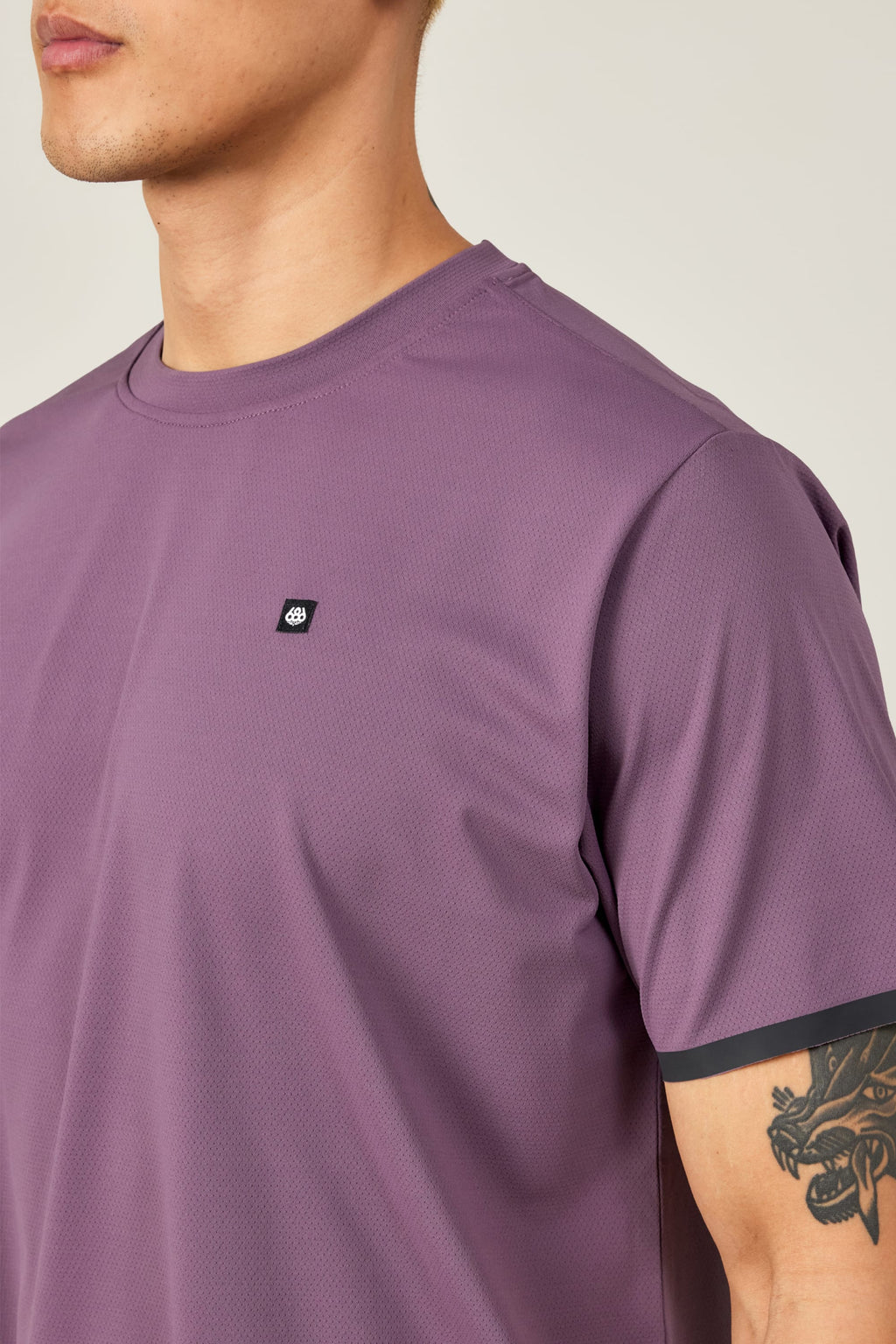 A person wears the 686 Men's Let's Go Tech Tee, a purple short-sleeve shirt with a small black and white 686 logo on the chest—ideal for spring-summer adventures. A black creature tattoo is visible on their upper right arm against a plain light background.