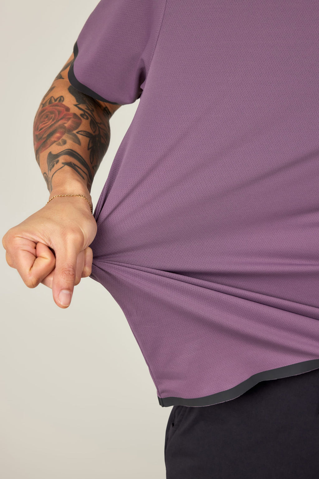 A tattooed person in a breathable 686 Men's Let's Go Tech Tee in purple pulls the fabric outward with their left hand, wearing a thin bracelet. The background is plain and light-colored.