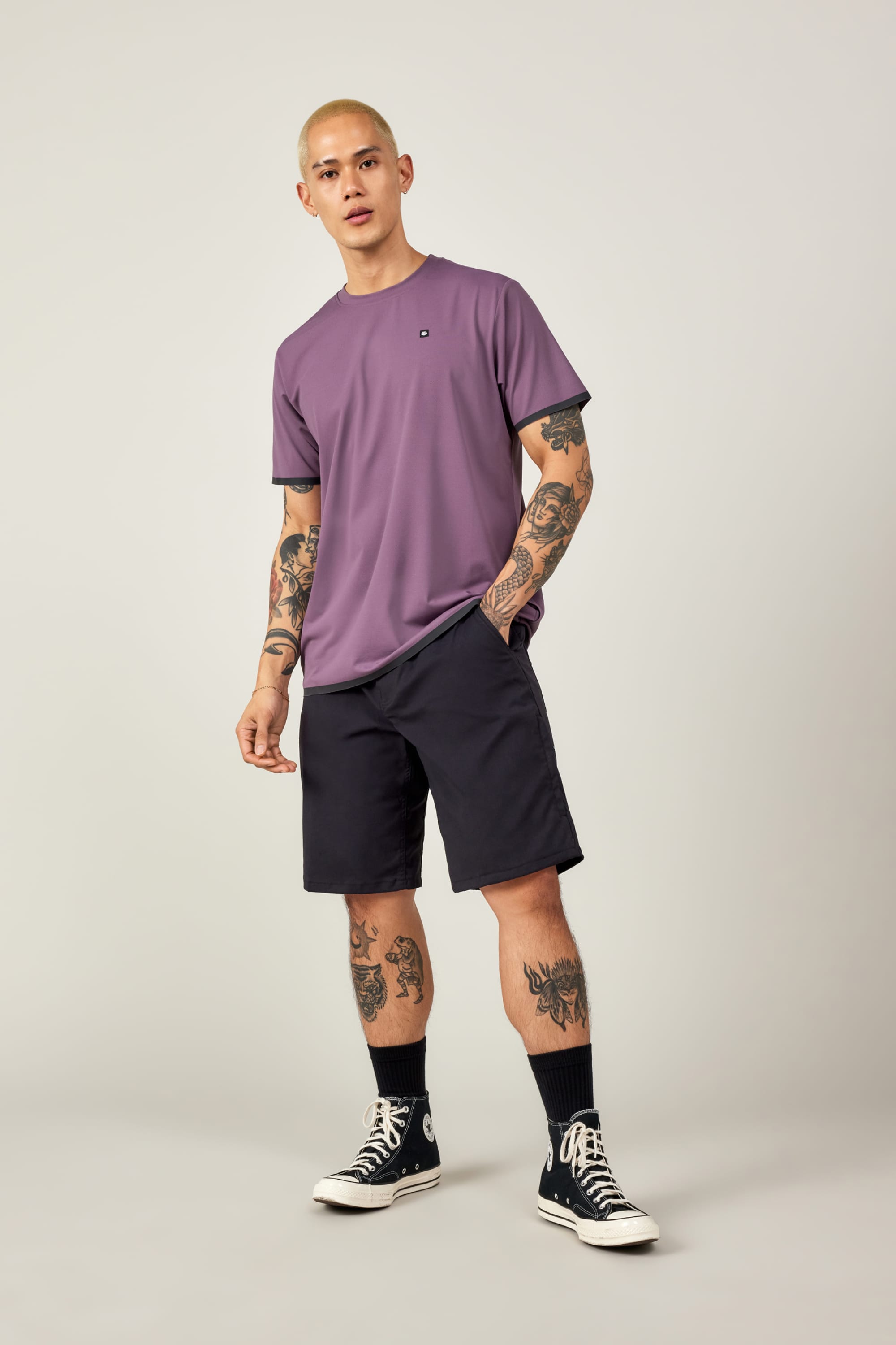 A person with short hair and tattoos wears the 686 Men's Let's Go Tech Tee in purple, black shorts, black socks, and black Converse sneakers, standing with one hand in their pocket against a plain light background.