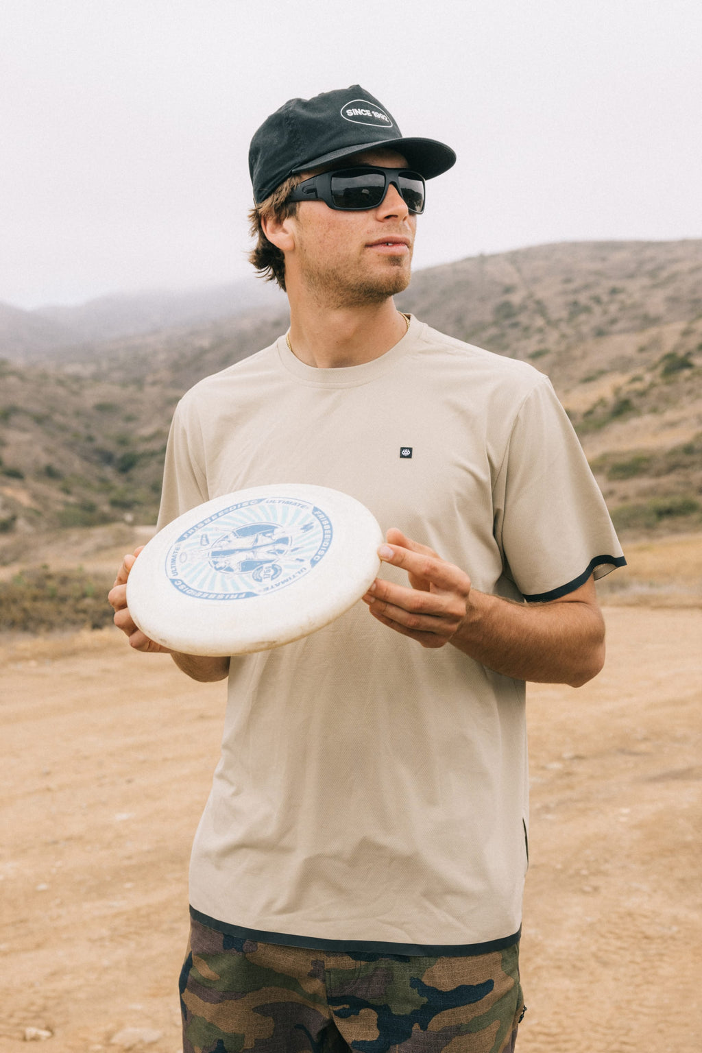 A man stands outdoors in a rugged landscape, wearing the 686 Men's Let's Go Tech Tee and sunglasses, holding a white frisbee—ready for spring and summer adventure under cloudy skies.
