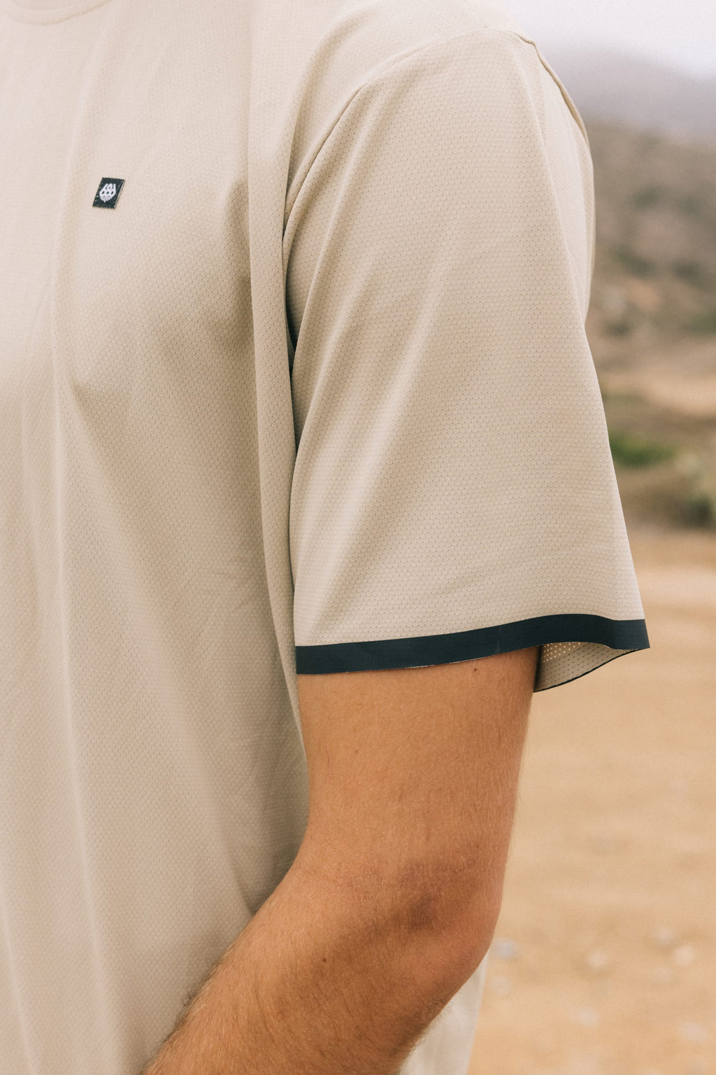 Wearing the 686 Men's Let's Go Tech Tee in beige with black-trimmed sleeves, a person stands outdoors—upper torso and arm shown against a blurred natural background—perfect for spring and summer adventures.