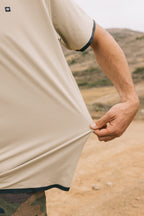 A person outdoors stretches the hem of a 686 Men's Let's Go Tech Tee with black trim; only one arm is visible and the scene shows a dirt path and hills, ideal for spring and summer adventures.