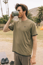 Wearing the 686 Men's Let's Go Tech Tee, a man sips from a green mug outdoors with palm trees, a tent, and hills in the background—perfect for a relaxed spring or summer adventure.