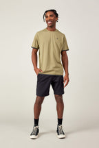 A man stands smiling against a plain background, wearing the 686 Men's Let's Go Tech Tee in olive green, black shorts, socks, and black-and-white sneakers. With one hand in his pocket, he looks relaxed and ready for any spring or summer adventure.