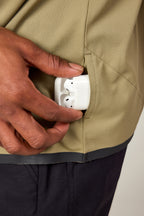 Someone slips a white wireless earbud case into a small zippered pocket on the olive green 686 Men's Let's Go Tech Tee—lightweight and ideal for spring or summer adventures.