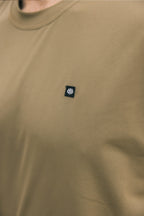 A close-up of a person wearing the 686 Men's Let's Go Tech Tee, a tan breathable shirt with tiny perforations and a black rectangular patch featuring the white 686 logo on the left chest—ideal for spring and summer adventures.