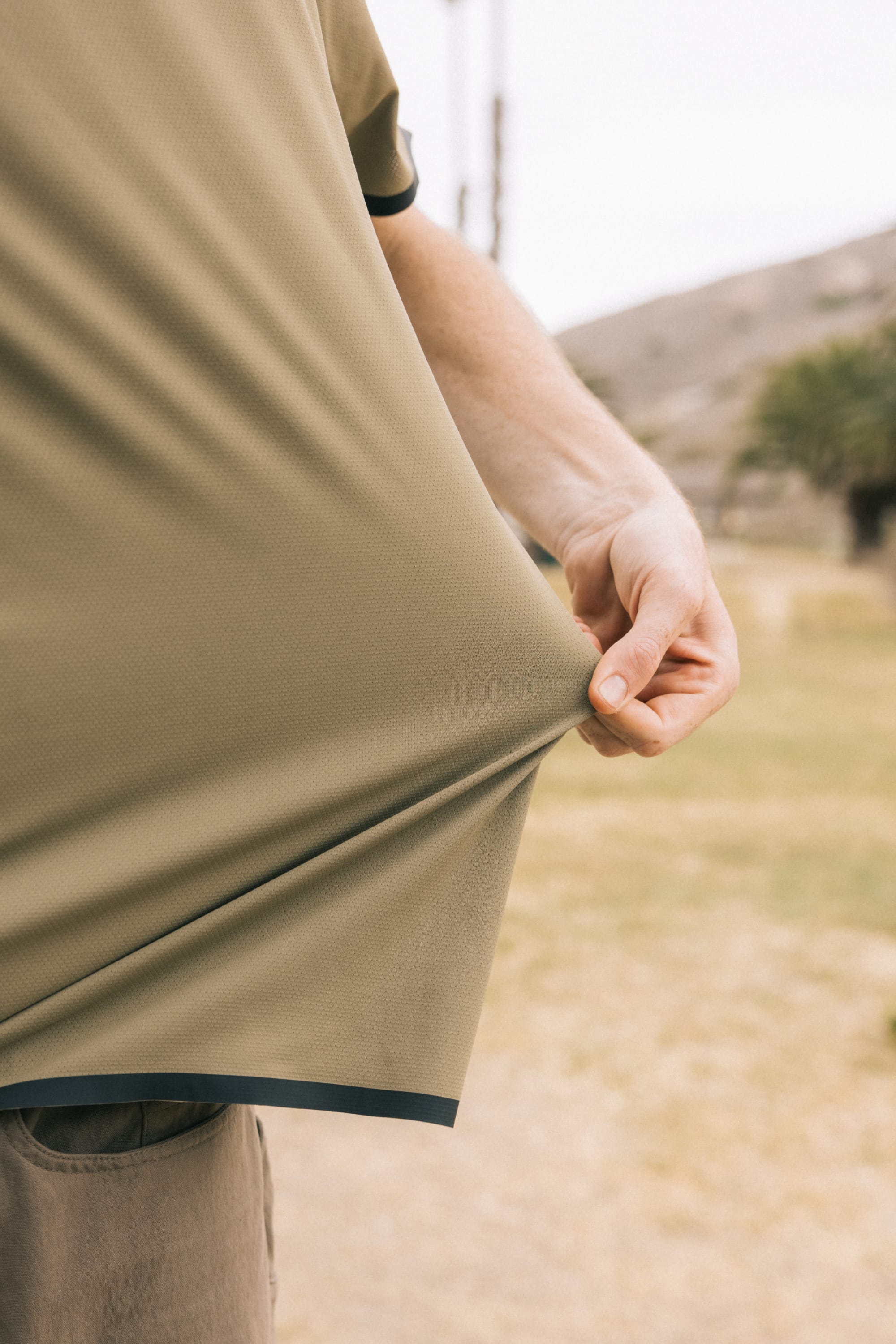 A person outdoors stretches the side of their light brown, breathable 686 Men's Let's Go Tech Tee. Only part of their torso and arm are visible, with grassy hills and blurred trees suggesting a spring or summer adventure.
