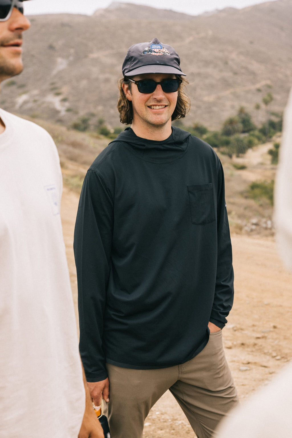 A man in sunglasses, a dark cap, and the 686 Men's Let's Go Tech Hoody stands outdoors on a dirt path with hills behind him. Two other people in light-colored shirts are partially visible nearby.