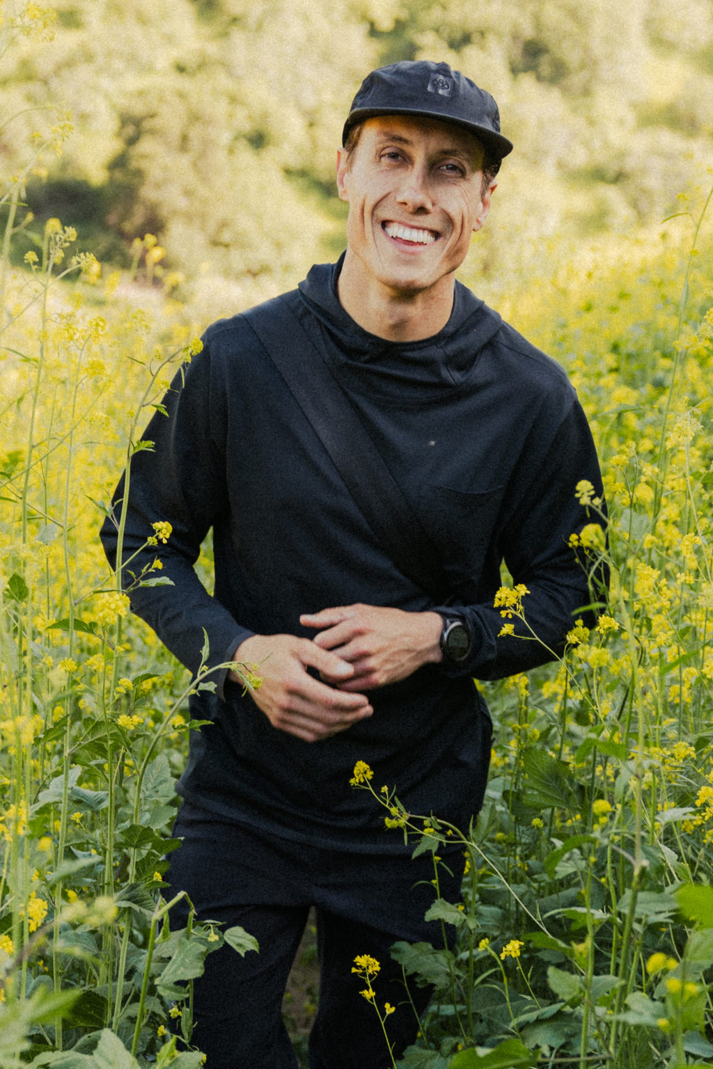 A man smiles in a field of yellow wildflowers, wearing the 686 Mens Lets Go Tech Hoody and a black cap, enjoying sun protection from his breathable 686 hoody on a warm day, with greenery and trees in the blurred background.