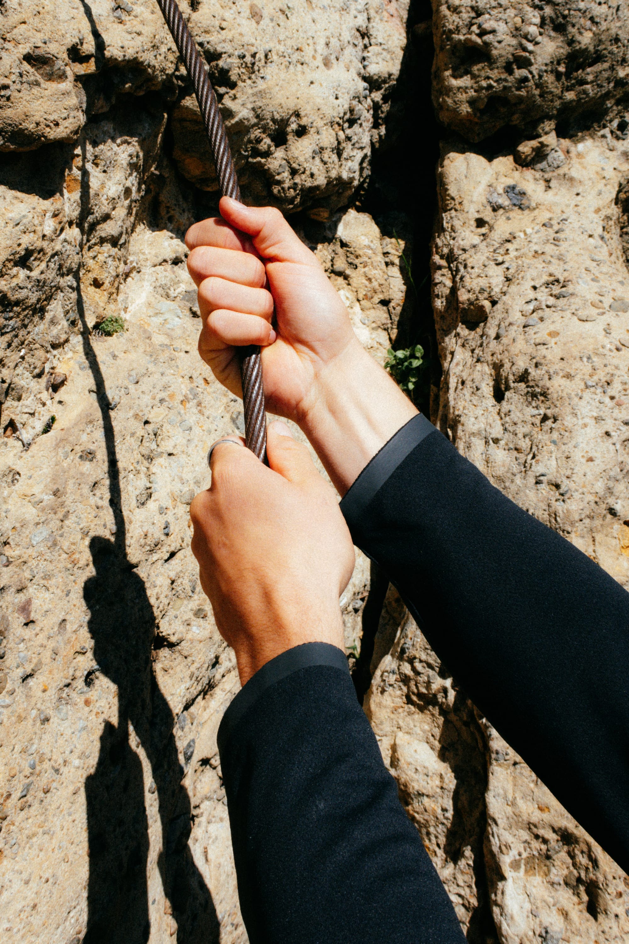 Two hands grip a thick rope against a rugged rock wall, suggesting climbing. The person wears the 686 Mens Lets Go Tech Hoody—a lightweight, breathable layer from 686 that offers sun protection as sunlight casts sharp shadows on the rocks.