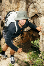 A young man in the 686 Mens Lets Go Tech Hoody climbs a rocky outdoor surface, wearing shorts and a hat, carrying a white backpack with lightweight gear as he looks up at the camera amid green plants.