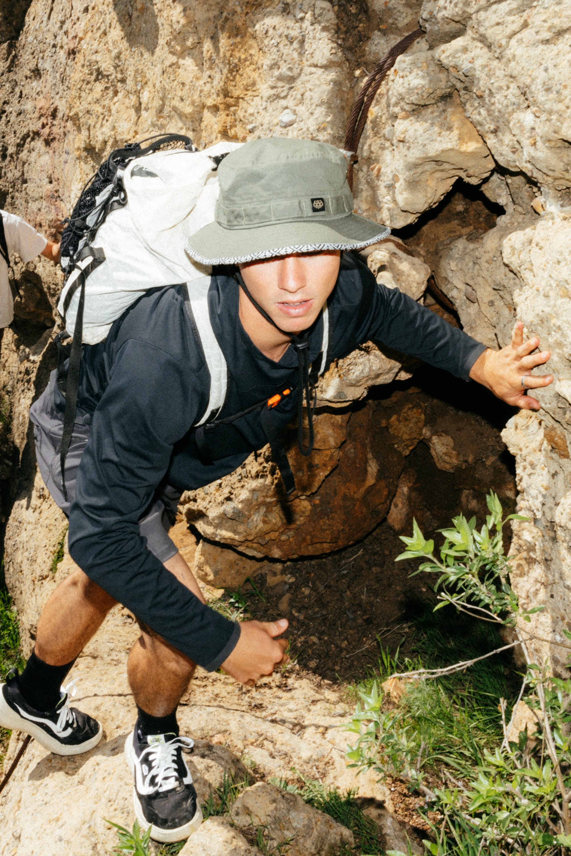 A young man in the 686 Mens Lets Go Tech Hoody climbs a rocky outdoor surface, wearing shorts and a hat, carrying a white backpack with lightweight gear as he looks up at the camera amid green plants.