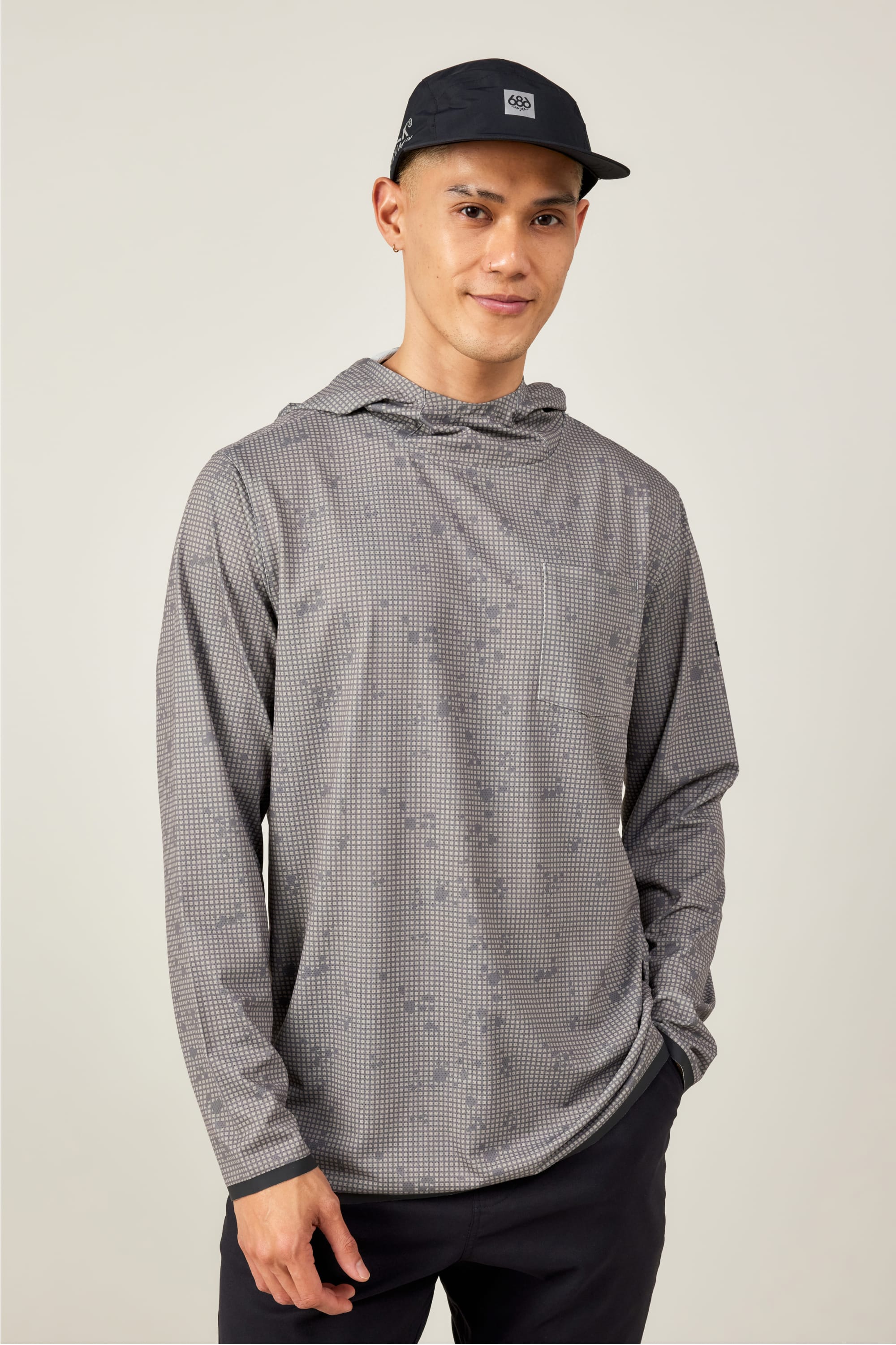 A person in a black baseball cap and the 686 Mens Lets Go Tech Hoody, featuring a lightweight gray patterned design, stands facing the camera with a slight smile and one hand in their pocket against a plain light background.