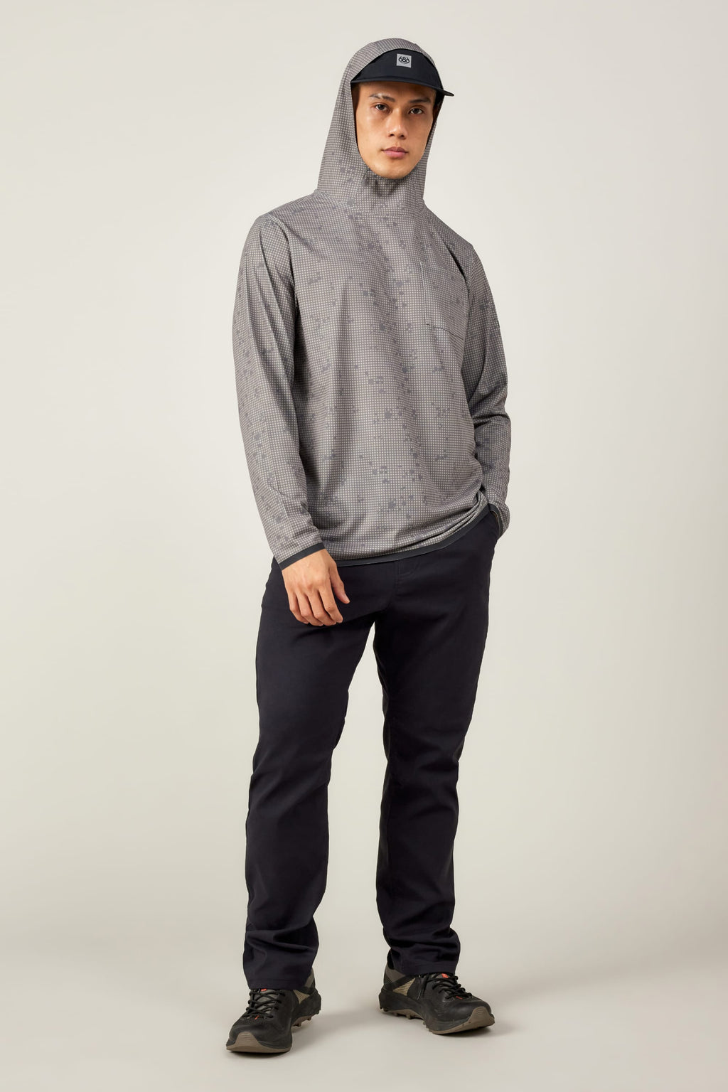 A person stands facing forward in the 686 Mens Lets Go Tech Hoody by 686—a lightweight, breathable top with a subtle pattern—paired with dark pants, black sneakers, and a gray cap, offering comfortable sun protection against a plain background.