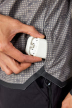 A person’s hand puts a white wireless earbud case into the zippered pocket of a lightweight, gray patterned 686 Mens Lets Go Tech Hoody.