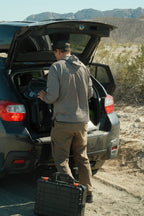 A man in the 686 Mens Lets Go Tech Hoody loads gear into an SUV on a dirt road, surrounded by desert and mountains.