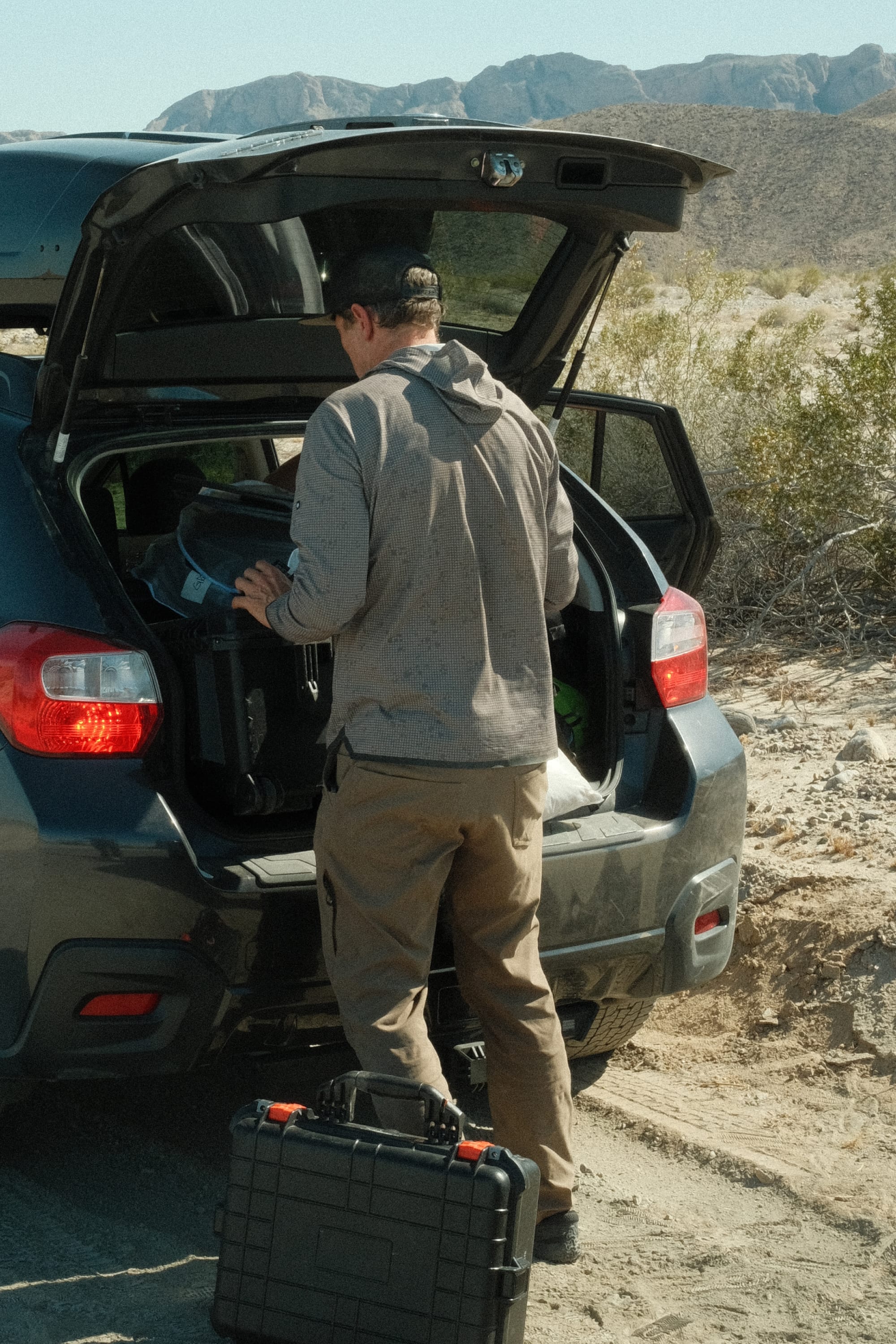 A man in the 686 Mens Lets Go Tech Hoody loads gear into an SUV on a dirt road, surrounded by desert and mountains.
