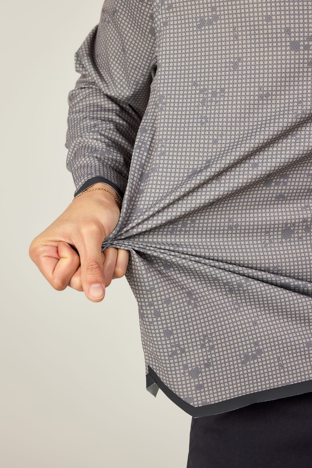 A person stretches the fabric of their gray, patterned 686 Mens Lets Go Tech Hoody with their left hand, highlighting its stretch and lightweight feel. This 686 top provides sun protection, shown against a plain light-colored background.