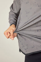 A person stretches the fabric of their gray, patterned 686 Mens Lets Go Tech Hoody with their left hand, highlighting its stretch and lightweight feel. This 686 top provides sun protection, shown against a plain light-colored background.