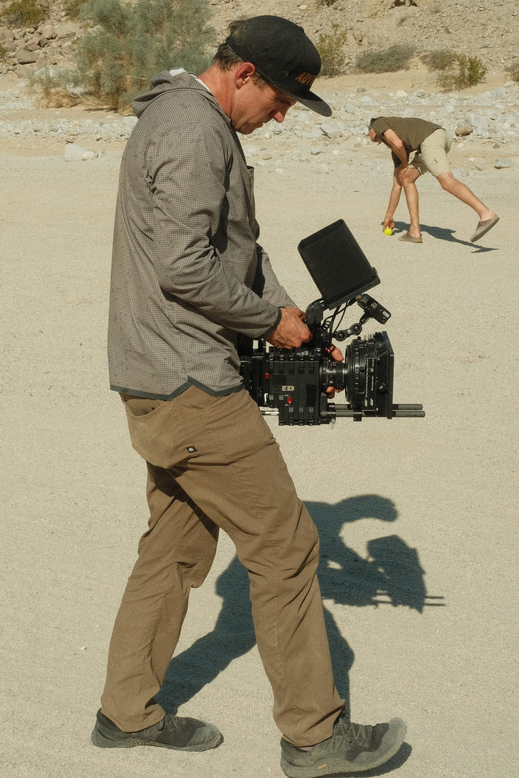A man wearing the 686 Mens Lets Go Tech Hoody by 686 operates a professional video camera outdoors on sandy ground, while another person in the background bends down to pick up something.