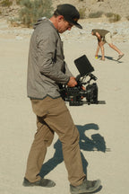 A man wearing the 686 Mens Lets Go Tech Hoody by 686 operates a professional video camera outdoors on sandy ground, while another person in the background bends down to pick up something.