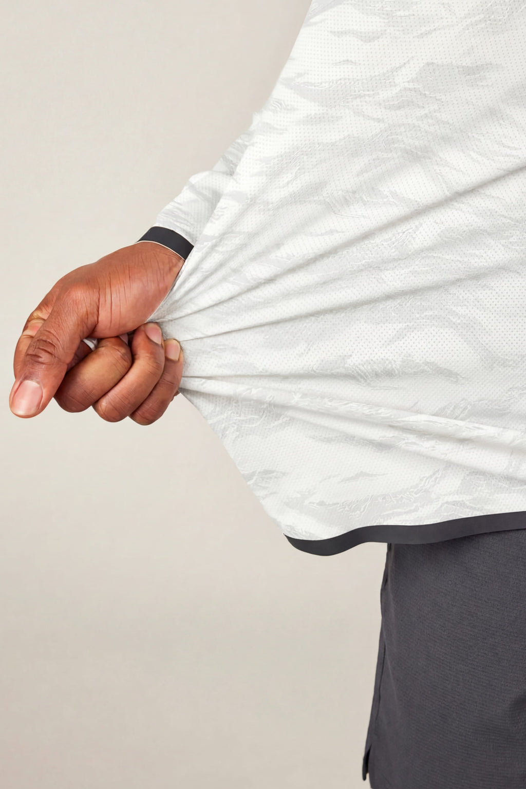 A hand grips and gently pulls the side of the 686 Men's Let's Go Tech Hoody in light gray, showing off the breathable, stretchy fabric. The person pairs it with dark pants and stands against a plain background.