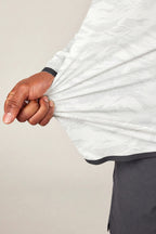 A hand grips and gently pulls the side of the 686 Men's Let's Go Tech Hoody in light gray, showing off the breathable, stretchy fabric. The person pairs it with dark pants and stands against a plain background.