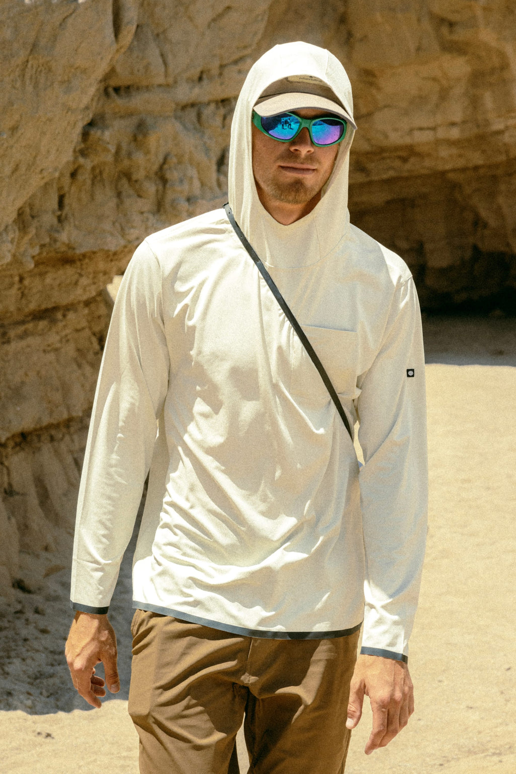 A man in the 686 Mens Lets Go Tech Hoody enjoys a sunny day outdoors, paired with brown pants, sunglasses, and a cap for added sun protection, with rocky cliffs in the background.