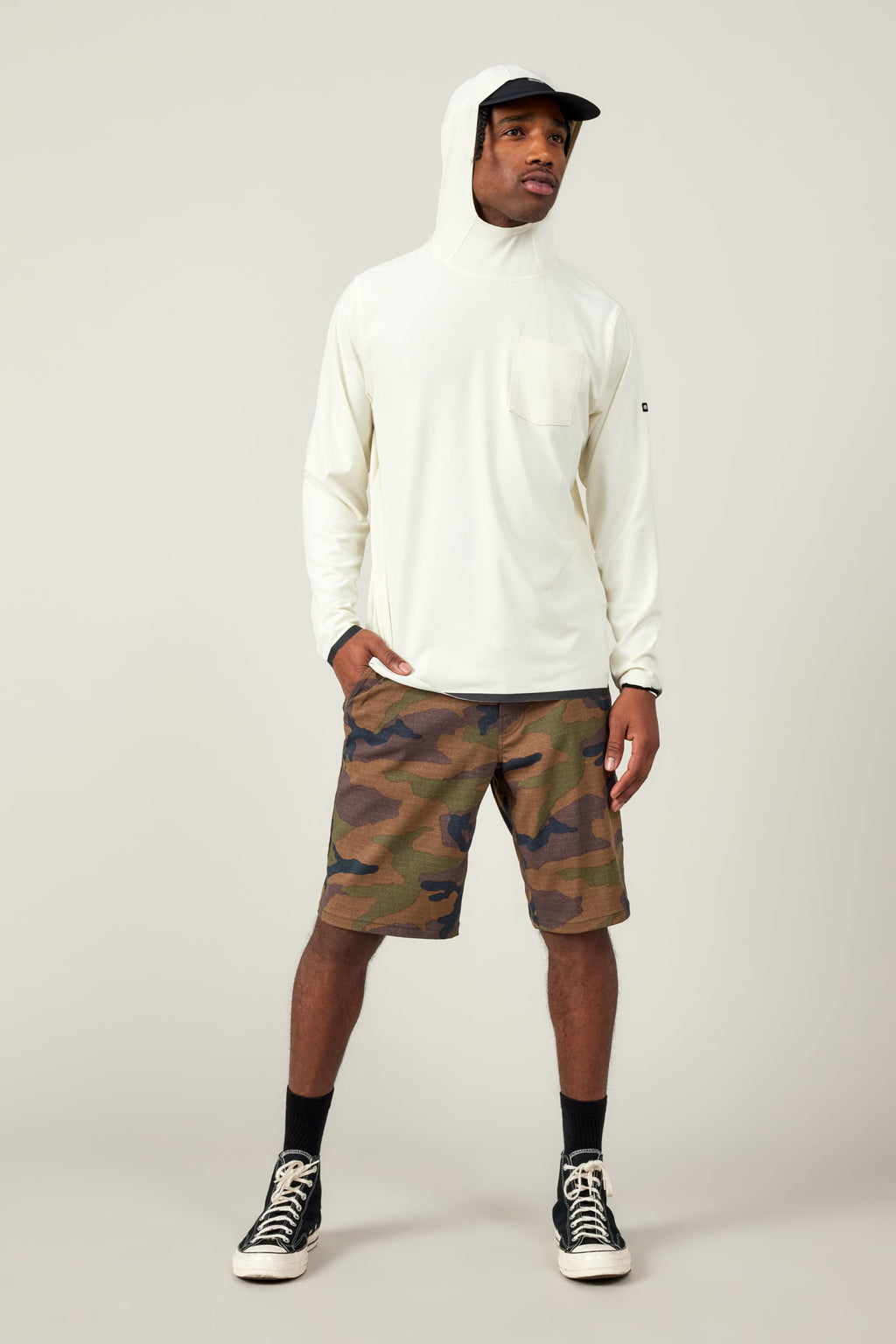 A man wearing the 686 Mens Lets Go Tech Hoody stands against a plain background, pairing it with a hat, camo shorts, black socks, and sneakers. He looks to the side with one hand in his pocket for added sun protection.