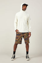 A man wearing the 686 Mens Lets Go Tech Hoody stands against a plain background, pairing it with a hat, camo shorts, black socks, and sneakers. He looks to the side with one hand in his pocket for added sun protection.