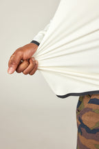 A person in camouflage pants stretches the side of their 686 Mens Lets Go Tech Hoody, a lightweight white long-sleeved shirt by 686, with their hand to show the fabric’s flexibility against a light background.