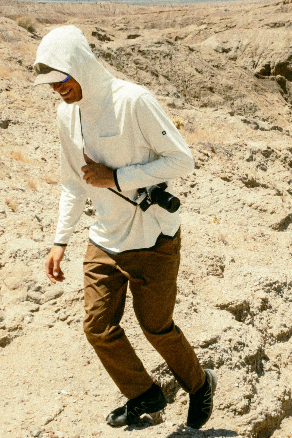 Wearing the 686 Mens Lets Go Tech Hoody, sunglasses, brown pants, and black shoes, a hiker treks across sunlit desert rocks with a camera over their shoulder for extra sun protection.
