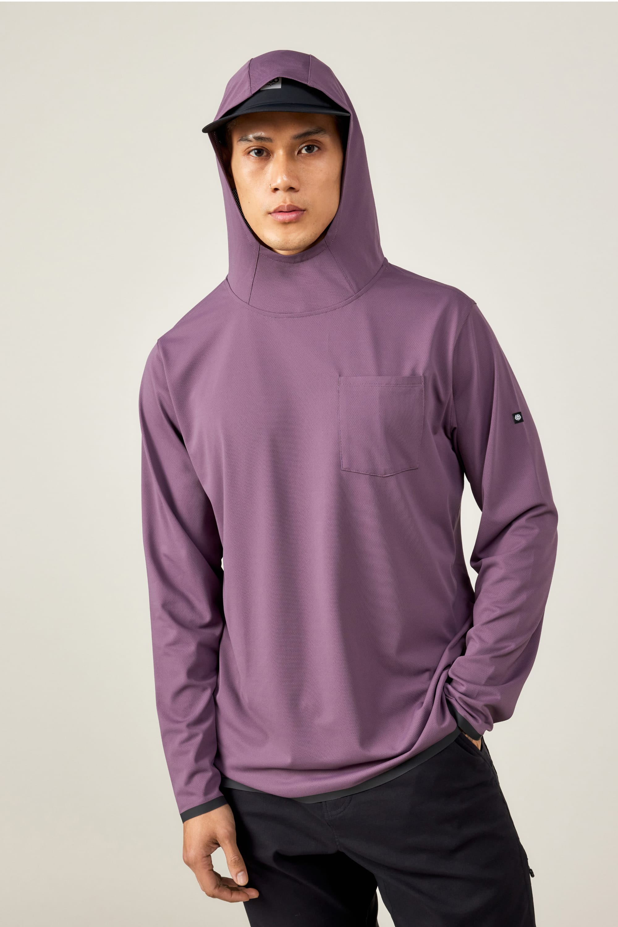 A person wearing the 686 Mens Lets Go Tech Hoody in purple with a chest pocket and hood, paired with a black cap, stands against a plain light background.