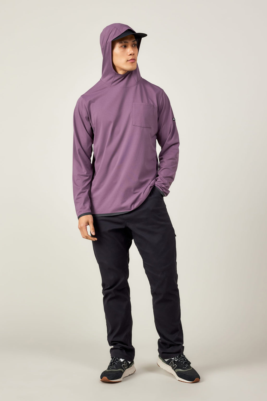 A person wears the 686 Mens Lets Go Tech Hoody in purple with a front pocket, paired with black pants, a black cap, and black sneakers, standing against a plain background and looking slightly to the side.