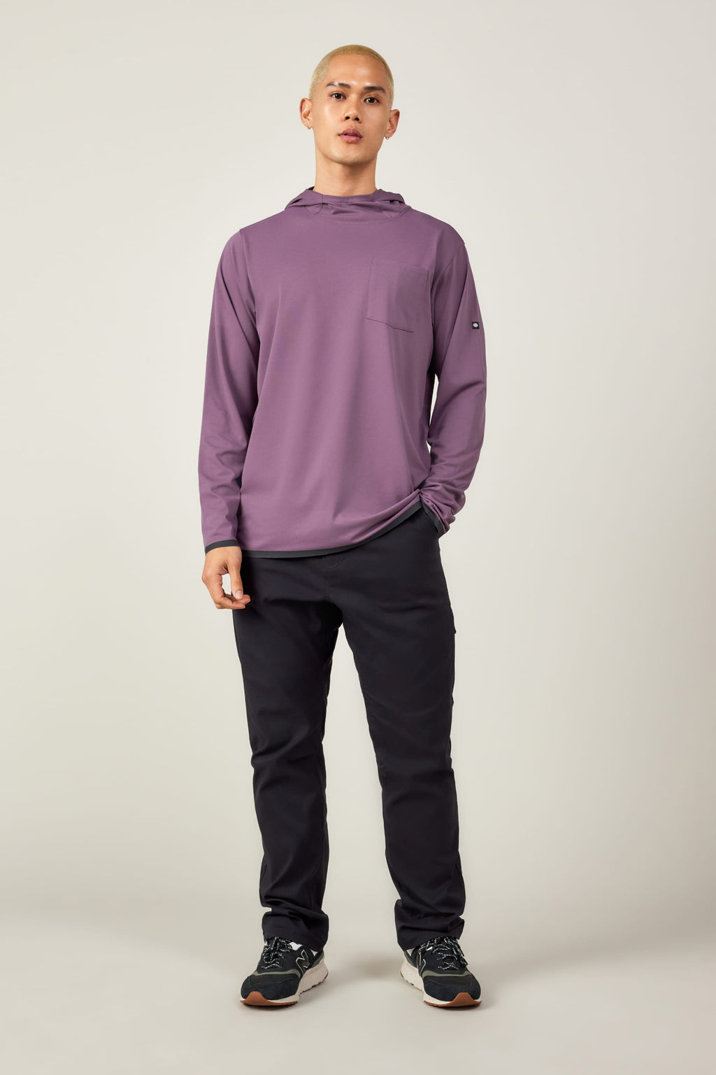 A person stands facing forward in a lightweight 686 Mens Lets Go Tech Hoody in light purple, paired with dark pants and black sneakers with white soles, set against a plain light background.