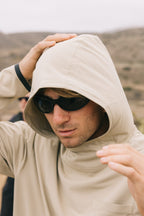 Outdoors against a blurred, natural backdrop, someone in the 686 Mens Lets Go Tech Hoody by 686 and dark sunglasses adjusts their hood.