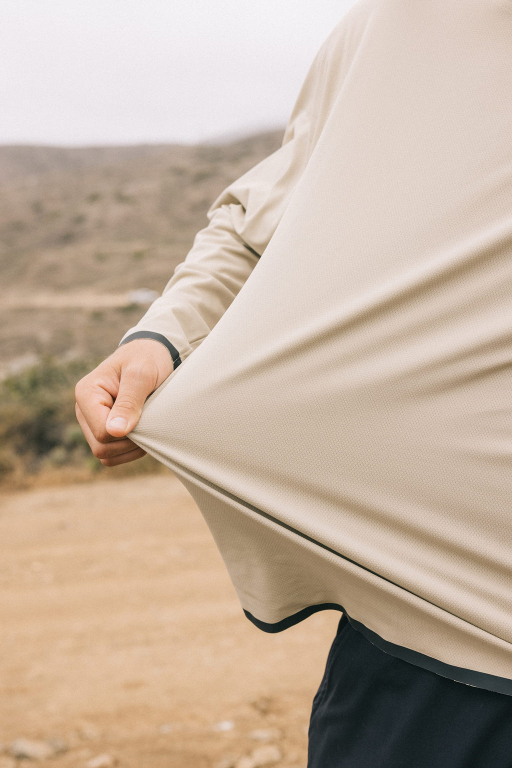 A person outdoors stretches the lightweight, beige 686 Mens Lets Go Tech Hoody away from their body. The background is a blurred, dry landscape with hills and scattered vegetation.