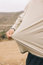 A person outdoors stretches the lightweight, beige 686 Mens Lets Go Tech Hoody away from their body. The background is a blurred, dry landscape with hills and scattered vegetation.