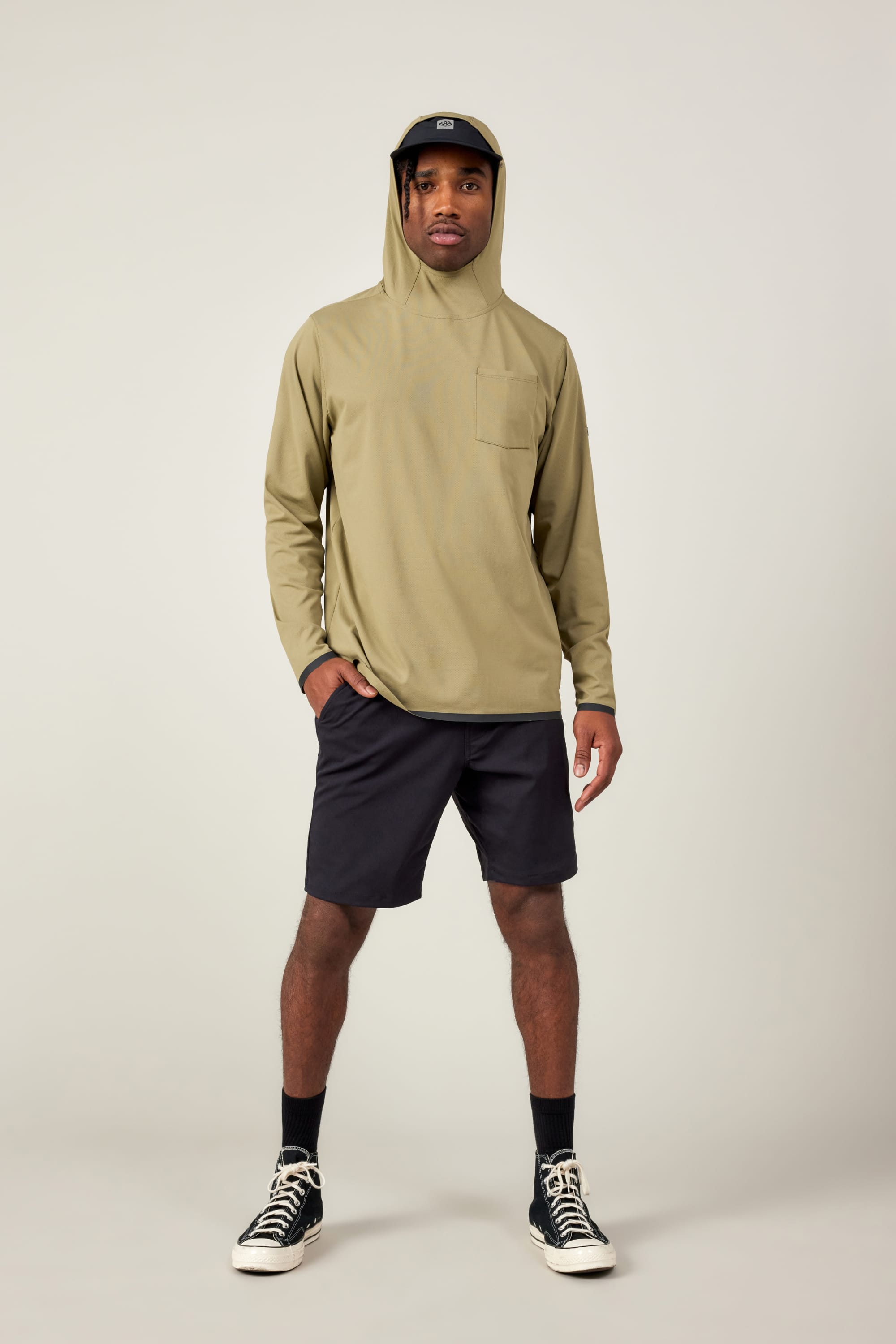 A man faces forward against a plain light background, wearing the 686 Mens Lets Go Tech Hoody in khaki for sun protection, paired with black shorts, socks, sneakers, and a dark visor cap.