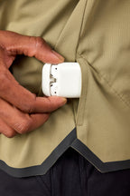 A person slides a white wireless earbud case into the side pocket of the 686 Mens Lets Go Tech Hoody by 686, featuring a lightweight, tan, textured design.