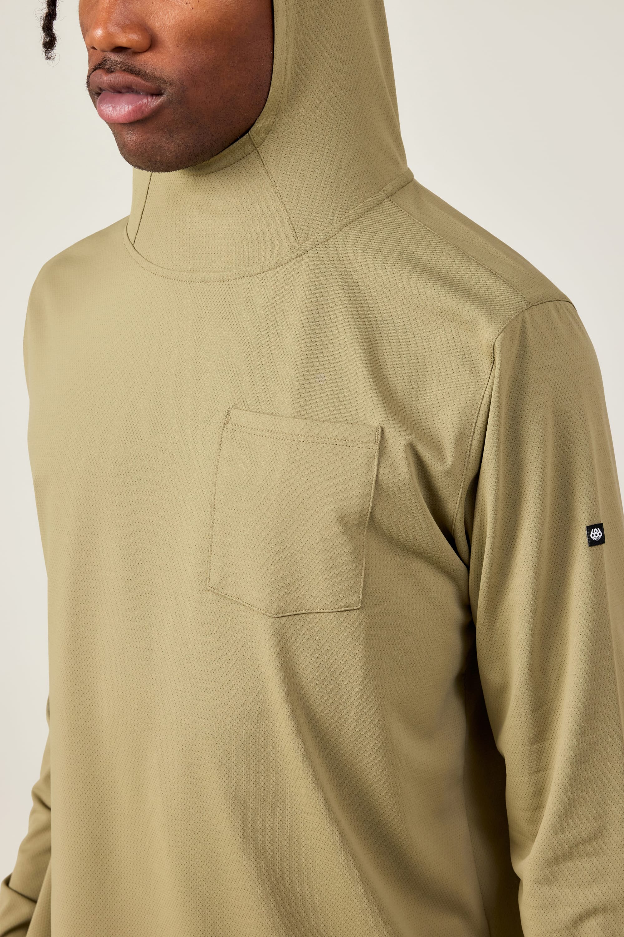 A person wearing the 686 Mens Lets Go Tech Hoody, a lightweight olive-green hoodie with a chest pocket and small black 686 logo patch on the sleeve. The image shows their upper body, with the face partially out of frame.